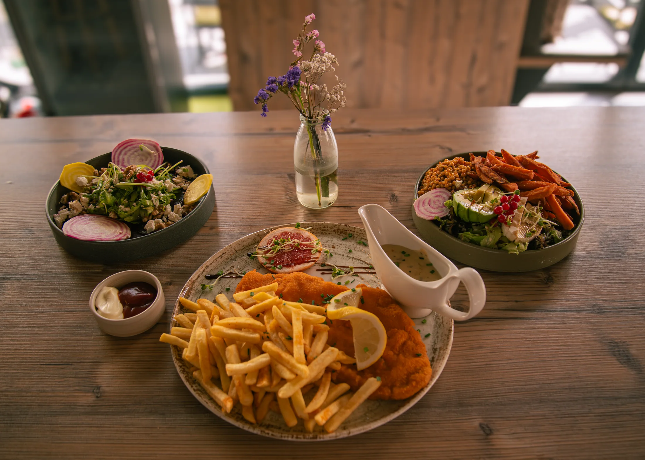 Zwei Bowls und ein klassisches paniertes Gericht mit Pommes stehen gemeinsam auf einem Holztisch.